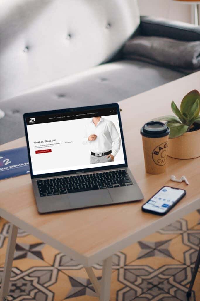 A laptop on a wooden table displays a website featuring a man in a white dress shirt. Nearby are a coffee cup, a smartphone, a plant, earphones, and a closed notebook. A gray sofa is in the background.